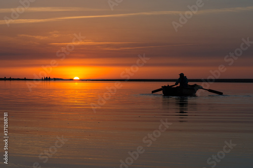 Young man fishing on a river from the wooden boat  at sunset . A fisherman in an old wooden boat rowing in the river at sunset © boulham