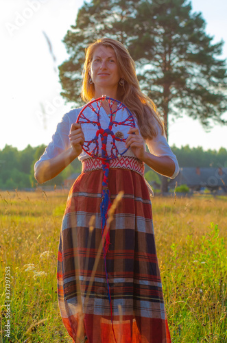 Beautiful romantic woman in a white blouse and long skirt with a symbol of a family tree on her hands against the background of a rural landscape at sunset. Summer nature, field and trees.  © Liudmila