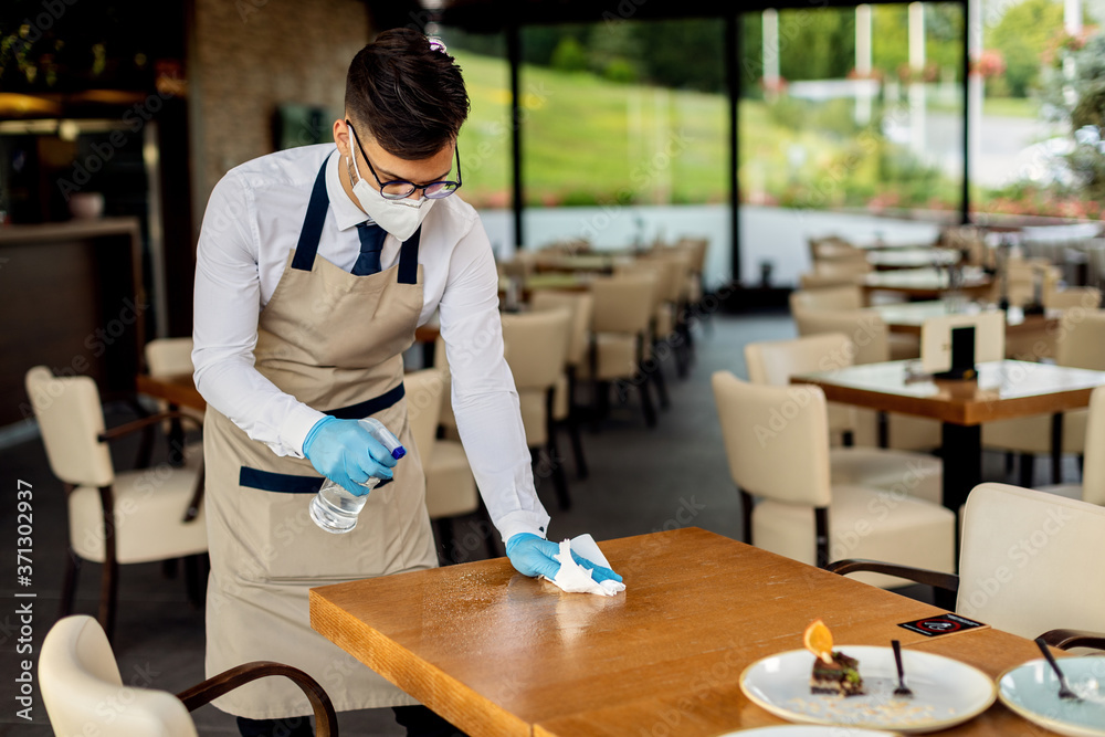 Waiter with protective face mask disinfecting tables in a cafe due to coronavirus epidemic.