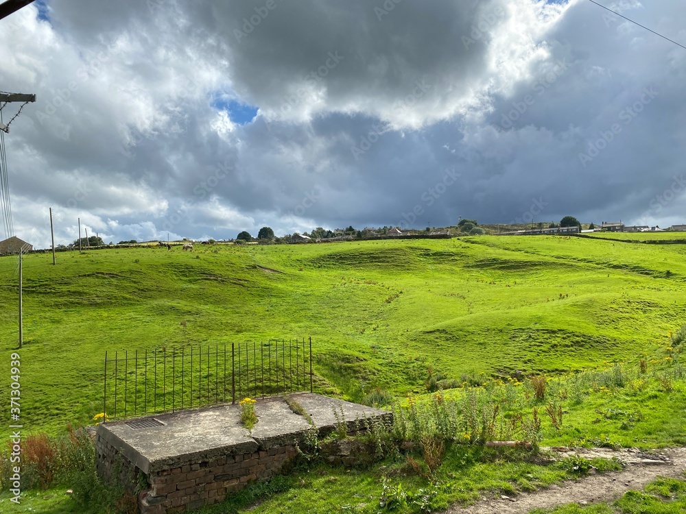 Heavy storm clouds over the Denholme countryside, with a green hillside, farm and houses
