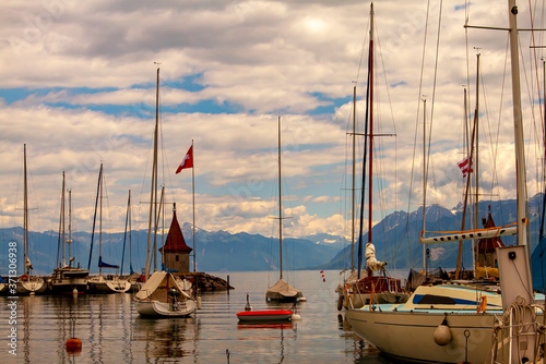A landscape view of the marina on the coast of Lake Geneva, Switzerland. Clear waters of the lake, a vintage lighthouse, dock, pier, sails and a Swiss flag is seen. Snowy Alps are seen across the lake