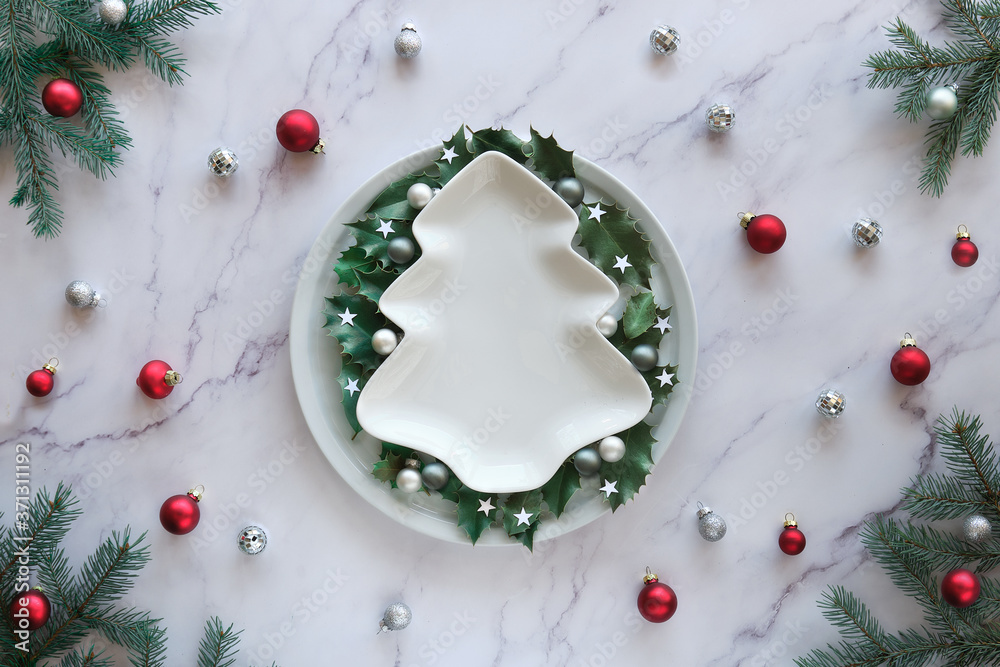 Christmas flat lay on marble table, copy-space on ceramic plate. Xmas background in green, white and red. Natural fir twigs, holly leaves and glass trinkets. Christmas flat lay on marble table, copy-space on ceramic plate. Xmas background in green, white and red. Natural fir twigs, holly leaves and glass trinkets.