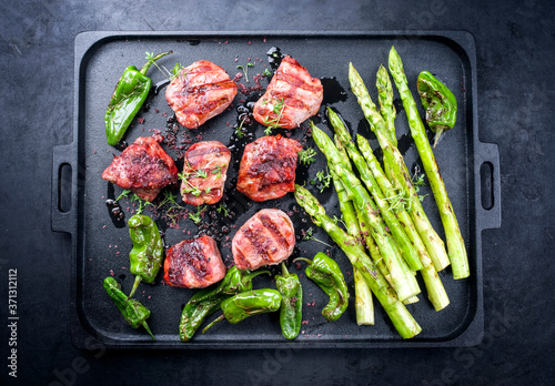 Traditional barbecue Iberian pork filet medaillons with green asparagus and chili offered as top view on a modern design cast iron black tray