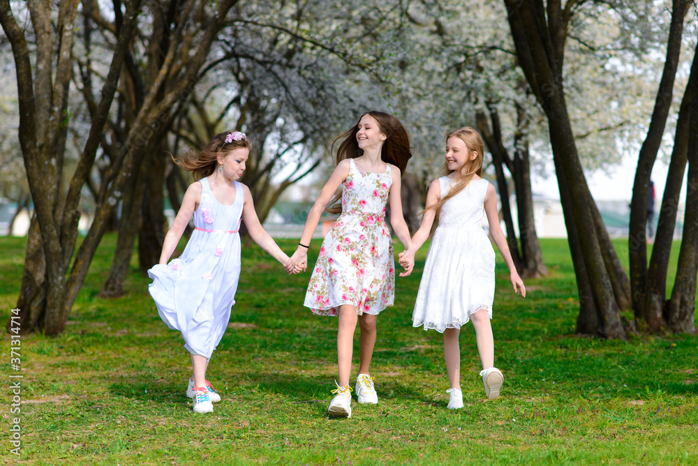 Three adorable girls in dresses hold hands in the garden, park.
