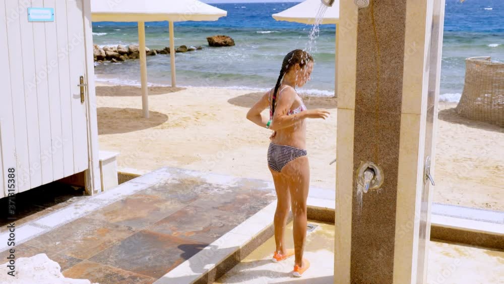 pretty teenage girl in a swimsuit, taking outdoor shower after sunbathing and swimming, on empty beach, by the sea. she rinses off the salt sea water, refreshing herself.