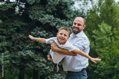 a bearded man rolls on the shoulders of a teenage boy in a park. Dad and son play airplanes in nature,