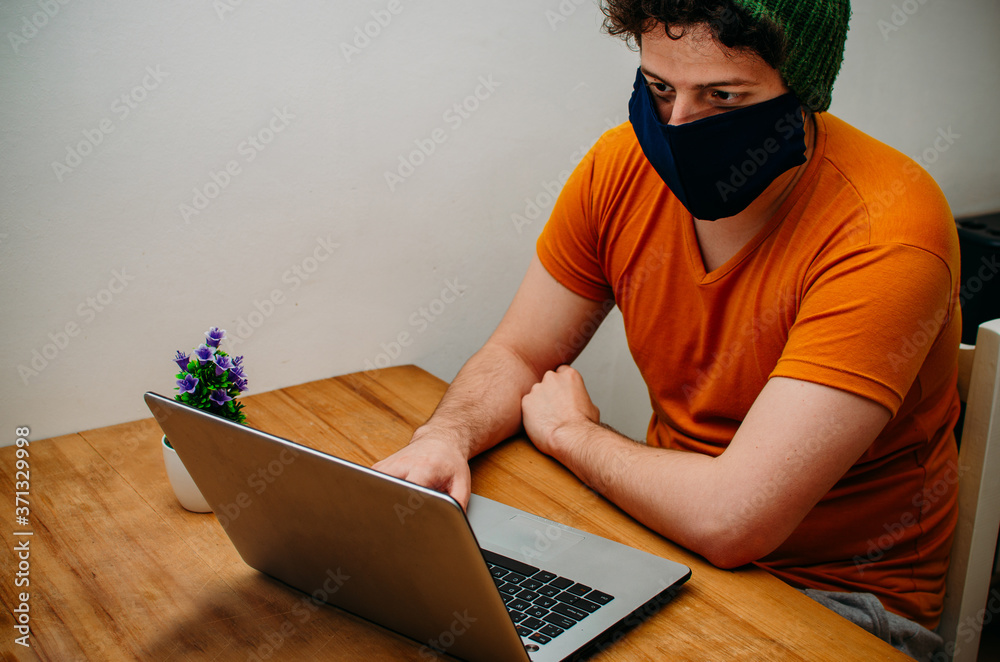 Joven de raza caucásica en el interior de su oficina trabajando en su portátil con mascarilla facial. Hombre con barbijo trabajando en su computadora personal sobre mesa de madera. Joven de raza caucásica en el interior de su oficina trabajando en su portátil con mascarilla facial. Hombre con barbijo trabajando en su computadora personal sobre mesa de madera.