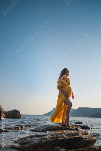 A beautiful pregnant woman in a yellow dress on the beach touching her belly with love and care. Walking on the seashore at sunset. Portrait on the background of the sea.