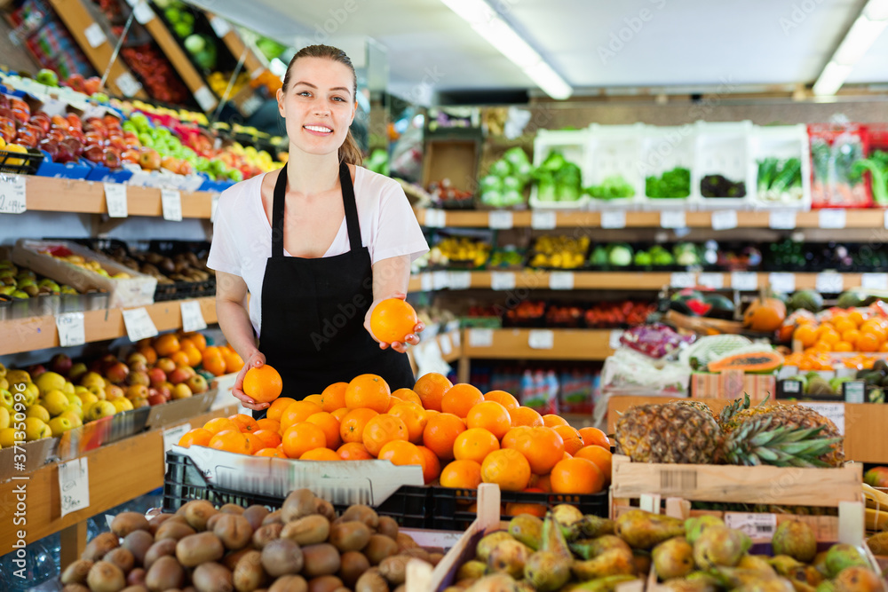 Young happy cheerful positive woman in apron working holding fresh oranges in hands on the supermarket