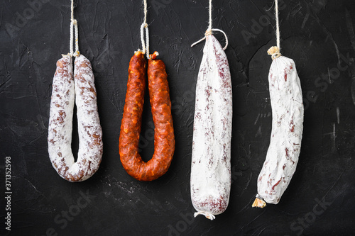 Spanish salami, fuet and salchichon sausages hang from a rack on black background, topview