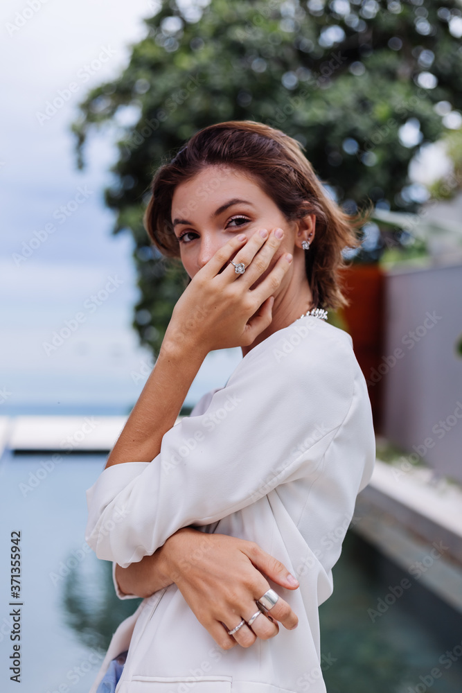 Fashion portrait of woman at tropical luxury villa wearing white stylish blazer, rings, earings on background of tropical leaves and swimming pool.