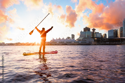 Adventurous man is paddle boarding near Downtown City during a vibrant winter sunrise. Taken in Coal Harbour, Vancouver, British Columbia, Canada. Colorful Sky Overlay