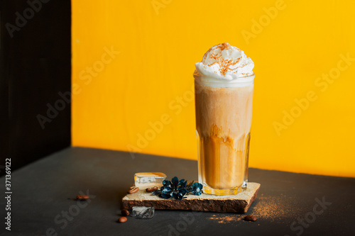 cold black coffee with whipped milk foam and spices in high glass on stand of natural stone, coffee beans and ice cubes on black table and yellow background