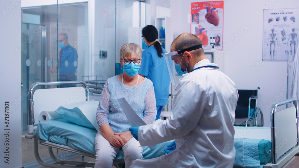 Obraz premium Medical stuff in visor explainingX Ray result to old retired woman sitting on hospital bed during COVID-19 pandemic. Nurse checking with old man in background. Modern private clinic or healthcare