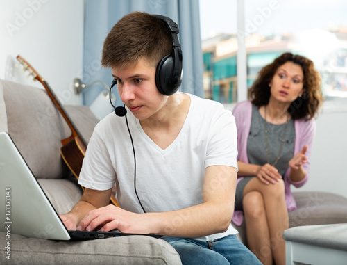 Teenage boy absorbedly playing on laptop at home on background of his worried mother.. photo