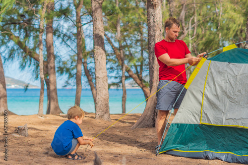 Camping people outdoor lifestyle tourists putting up setting up their green grey campsite in summer forest near lazur sea. Cute boy son helps his father assembling tent. Natural education of children