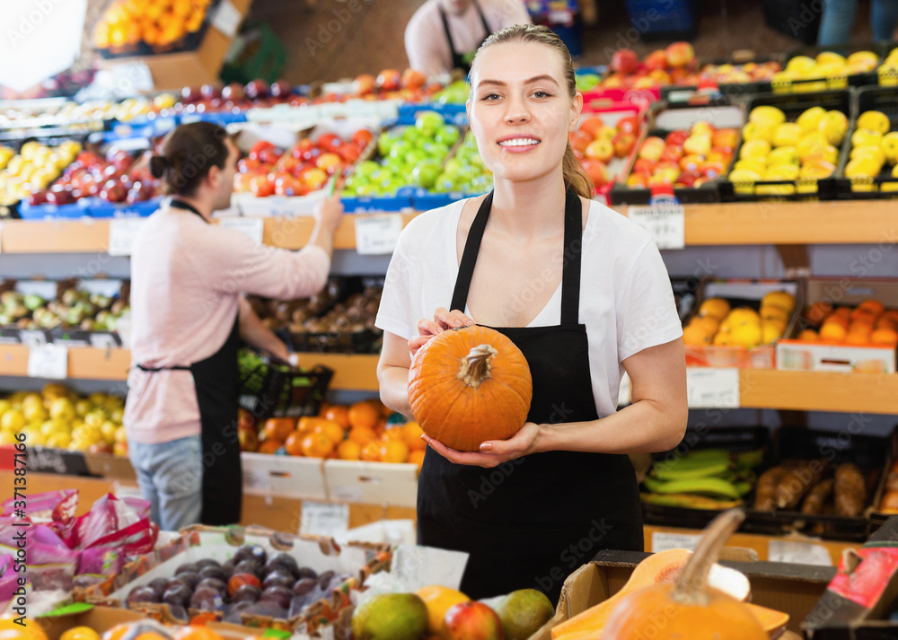 Obraz premium Young satisfied pleasant smiling woman with pumpkin in hands and man on background on the supermarket
