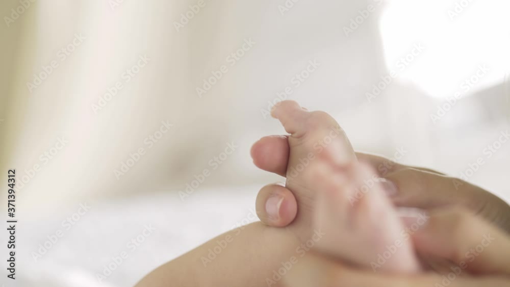 smiling mother runs fingers on little baby bare feet and toes practicing massage at home against blurry white wall under bright sunlight closeup smiling mother runs fingers on little baby bare feet and toes practicing massage at home against blurry white wall under bright sunlight closeup