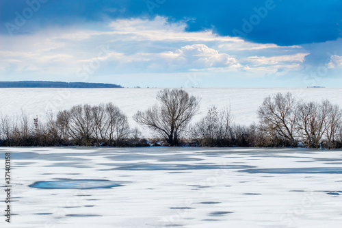 Winter landscape with river, trees, field and dramatic sky in sunny weather