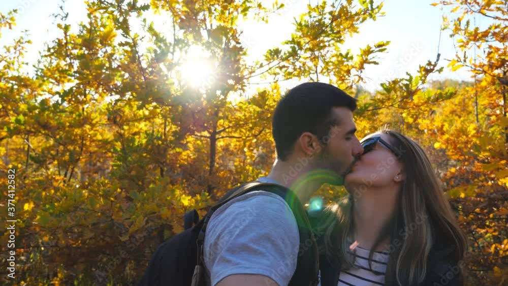 Portrait of happy smiling couple kisses while takes selfie photo with autumn garden at background. Young cheerful lovers stands at nature and looks into camera. Concept of loving. Slow motion Portrait of happy smiling couple kisses while takes selfie photo with autumn garden at background. Young cheerful lovers stands at nature and looks into camera. Concept of loving. Slow motion