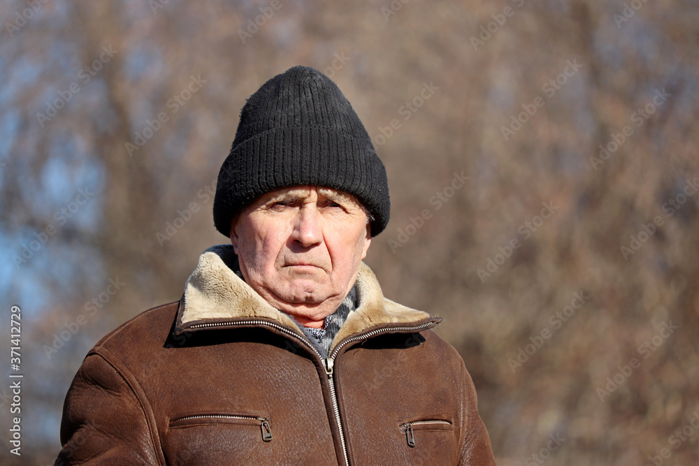 Portrait of elderly man with stern face standing on autumn rural background. Concept of cold weather, life in village, old age