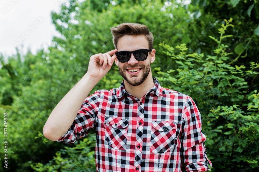 Perfect male. young unshaven man. sexy man wearing checkered clothes posing in park. Good confident young man standing and smiling in urban shirt. denim fashion style. handsome man with beard Perfect male. young unshaven man. sexy man wearing checkered clothes posing in park. Good confident young man standing and smiling in urban shirt. denim fashion style. handsome man with beard