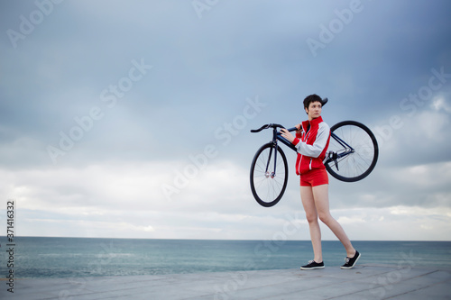 Full length portrait of professional female rider dressed in red sport jacket and shorts holding her light weight fixed gear bicycle while standing on concrete pier seashore with copy space cloudy sky photo