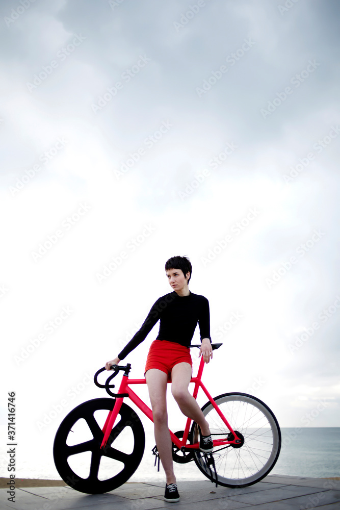Obraz premium Portrait of professional female cyclist enjoying the rest after bike ride along seashore while standing on the pier against sea and sky background with copy space area for your text message or content