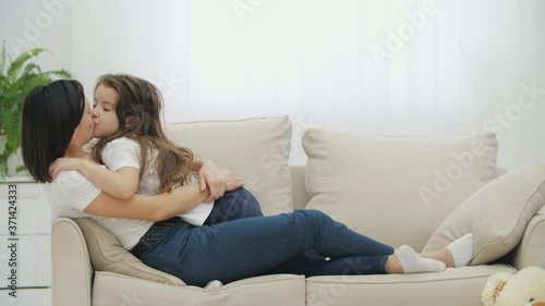Cute little kid girl and lovely mother are lying on the sofa, smiling, kissing, and hugging each other.