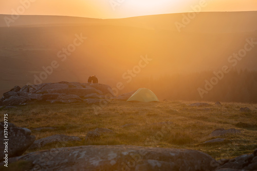 Stunning image of unidentified young couple wild camping in English countryside watching the stunning Summer sunrise with warm glow of the sun lighting the landscape