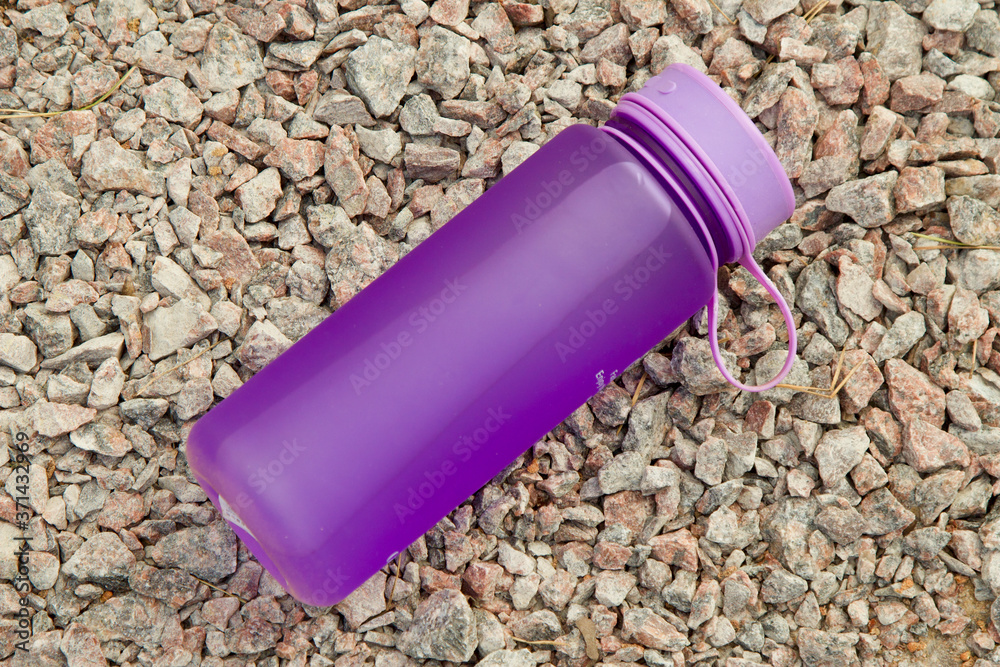 Top view of a colored reusable plastic lilac bottle for water close-up on a rocky background. The concept of sports, ecology, tourism and outdoor activities. Top view of a colored reusable plastic lilac bottle for water close-up on a rocky background. The concept of sports, ecology, tourism and outdoor activities.