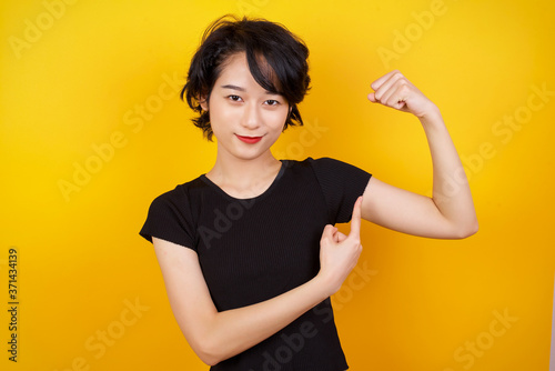 Waist up shot of blonde woman raises hand to show her muscles, feels confident in victory, looks strong and independent, smiles positively at camera, stands against gray background. Sport concept. photo