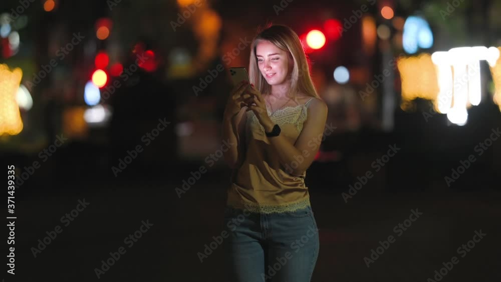 Cute Girl Uses Smartphone and Smiles Standing on Night Street in the City. Woman Messaging on Phone at Night on the Street With the Lights of the Road Blurred Background. Cute Girl Uses Smartphone and Smiles Standing on Night Street in the City. Woman Messaging on Phone at Night on the Street With the Lights of the Road Blurred Background.