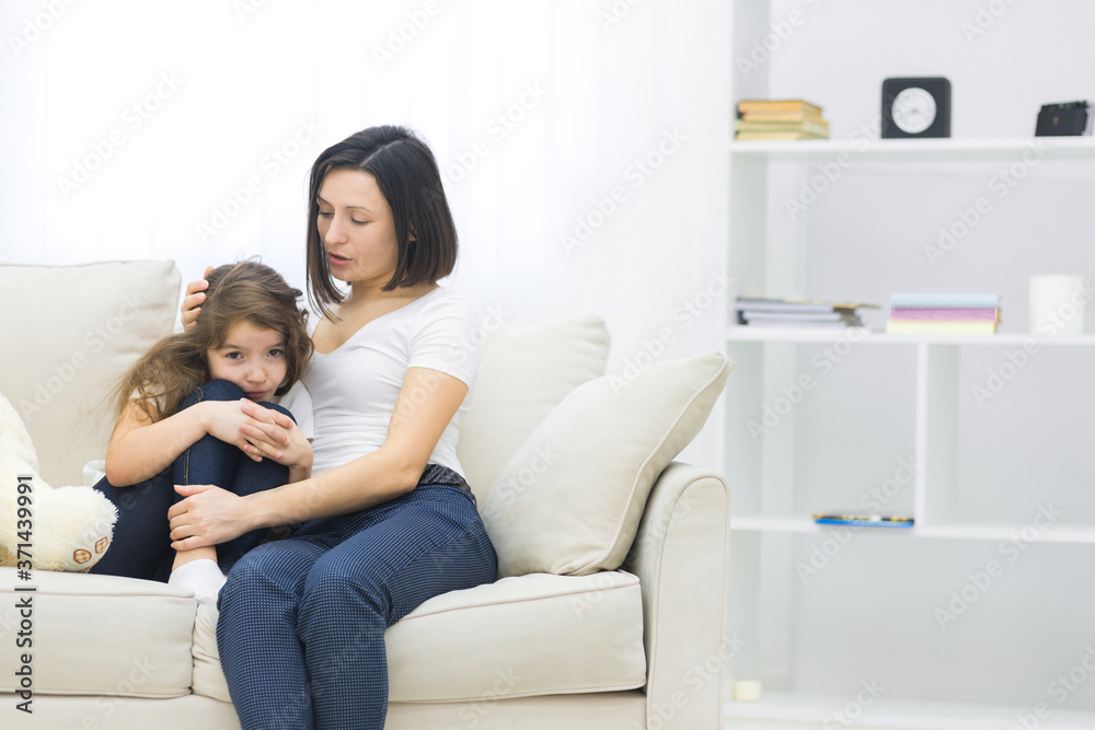 Photo of sad little girl sitting on the sofa and her mom hugging her.
