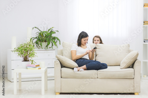 Photo of mother sitting on white sofa in her phone and her daughter hiding behind the sofa.