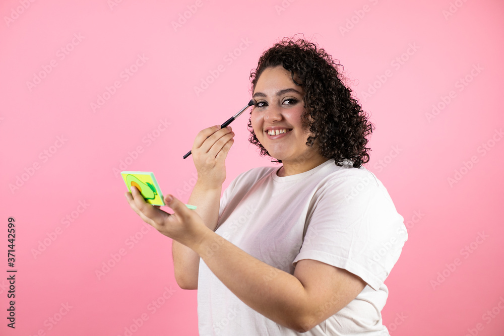 Young beautiful woman standing over isolated pink background smiling, looking in a mirror and putting on makeup Young beautiful woman standing over isolated pink background smiling, looking in a mirror and putting on makeup
