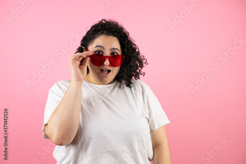 Young beautiful woman wearing sunglasses over isolated pink background surprised and taking and lowering her sunglasses © Irene