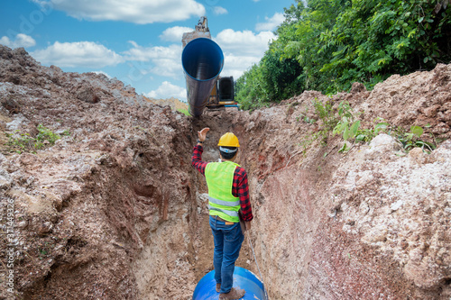 Engineer wear safety uniform hold a laptop examining excavation Drainage Pipe and Large plumbing water system underground at construction site.
