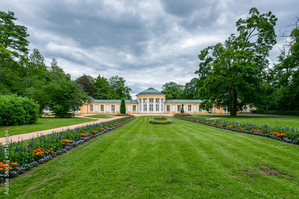 Marianske Lazne / Czech Republic - July 12 2020: View of the Ferdinand pavilion standing in a park with trees, green lawn and colorful flowers on a summer day. Grey sky.