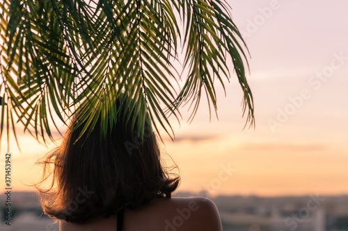 Back view of a beautiful girl in a straw hat against the background of the sea in branches of palm trees. Sunset beach. Summer and freedom concept.