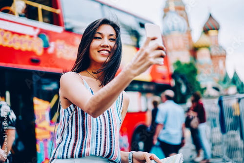 Cheerful female traveler making photo on smartphone camera during city tour standing near double decker,smiling asian girl taking selfie for share in networks having trip on sightseeing bus