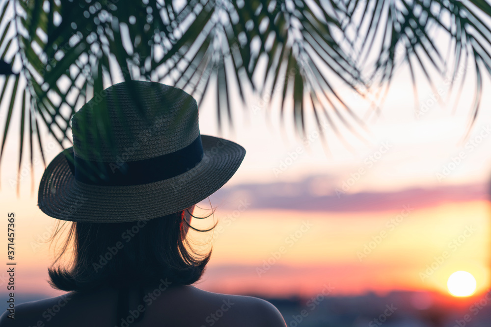 Back view of a beautiful girl in a straw hat against the background of the sea in branches of palm trees. Sunset beach. Summer and freedom concept.