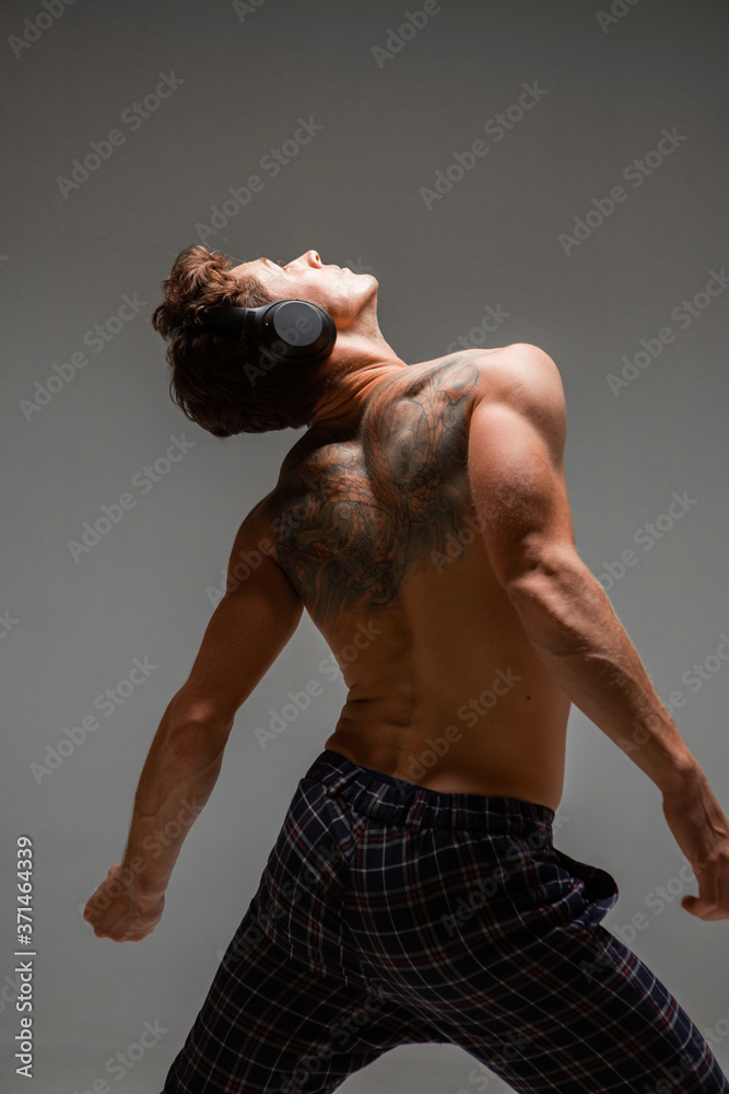 Cool young guy in wireless headphone listen to song with naked back isolated on gray background. Body with tattoo