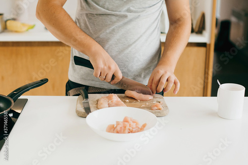 Person cutting chicken fillet with knife on wooden board on modern white kitchen. Process of making home pizza, hands close up. Home cooking concept
