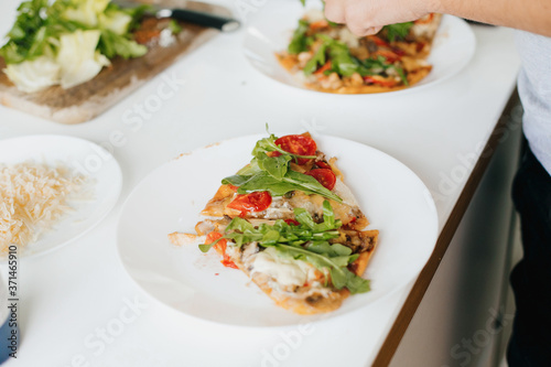 Slice of delicious pizza with mushrooms  tomato  arugula and cheese. Home cooking concept. Hands putting parmesan slices on homemade pizza on plate on modern kitchen.