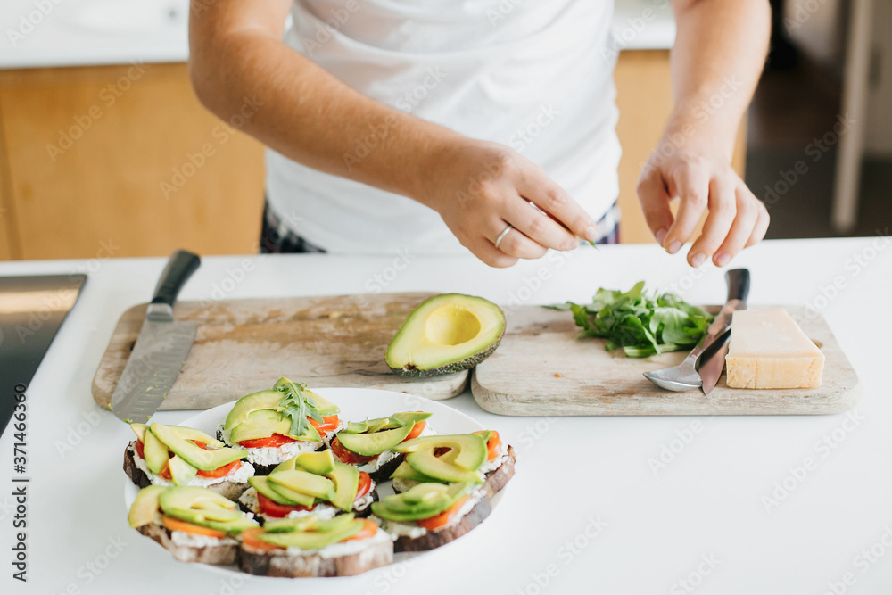 Process of making toasts with avocado on modern white kitchen. Hand putting arugula on sandwich made of whole grain bread, avocado, tomato, and cheese. Home cooking concept