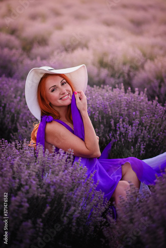 Happy young red-haired woman sitting in lavender field at sunset