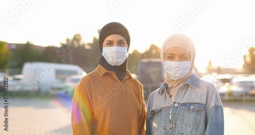 Portrait of beautiful happy Arabian women in traditional headscarves and medical masks looking at camera in sunlight. Sunny summer outdoor. Muslim cheerful females in hijabs. Islamic girls in city. photo