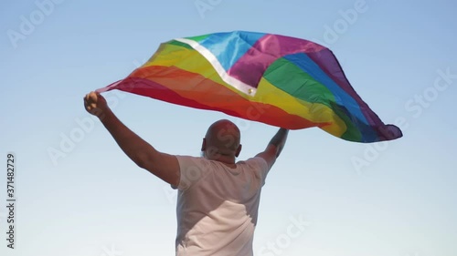 Bold man with rainbow gay flag while parade on background of blue sky. LGBTQI, Pride Event, LBGT Pride Month, Gay Pride Symbol