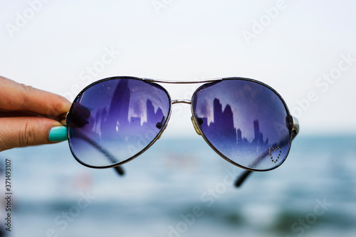 Female hand holds colorful sunglasses against the background of the sky and blue sea. The points reflect the storey buildings of the city © Venera